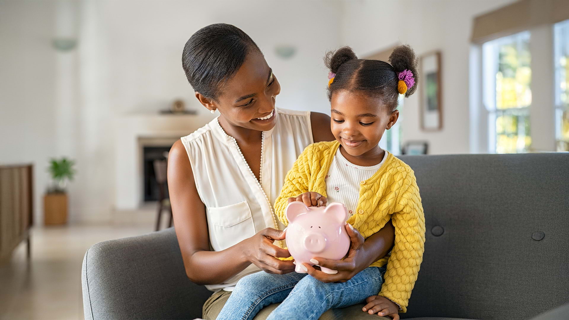 Smiling mother helping daughter sitting on lap putting money in piggy bank.